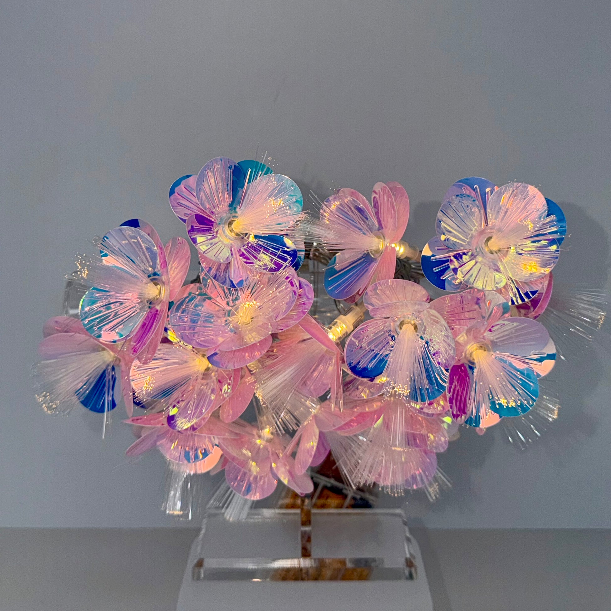 A decorative floral arrangement made of translucent iridescent flower petals with glowing warm lights inside, mounted on a clear acrylic stand against a plain light gray background.