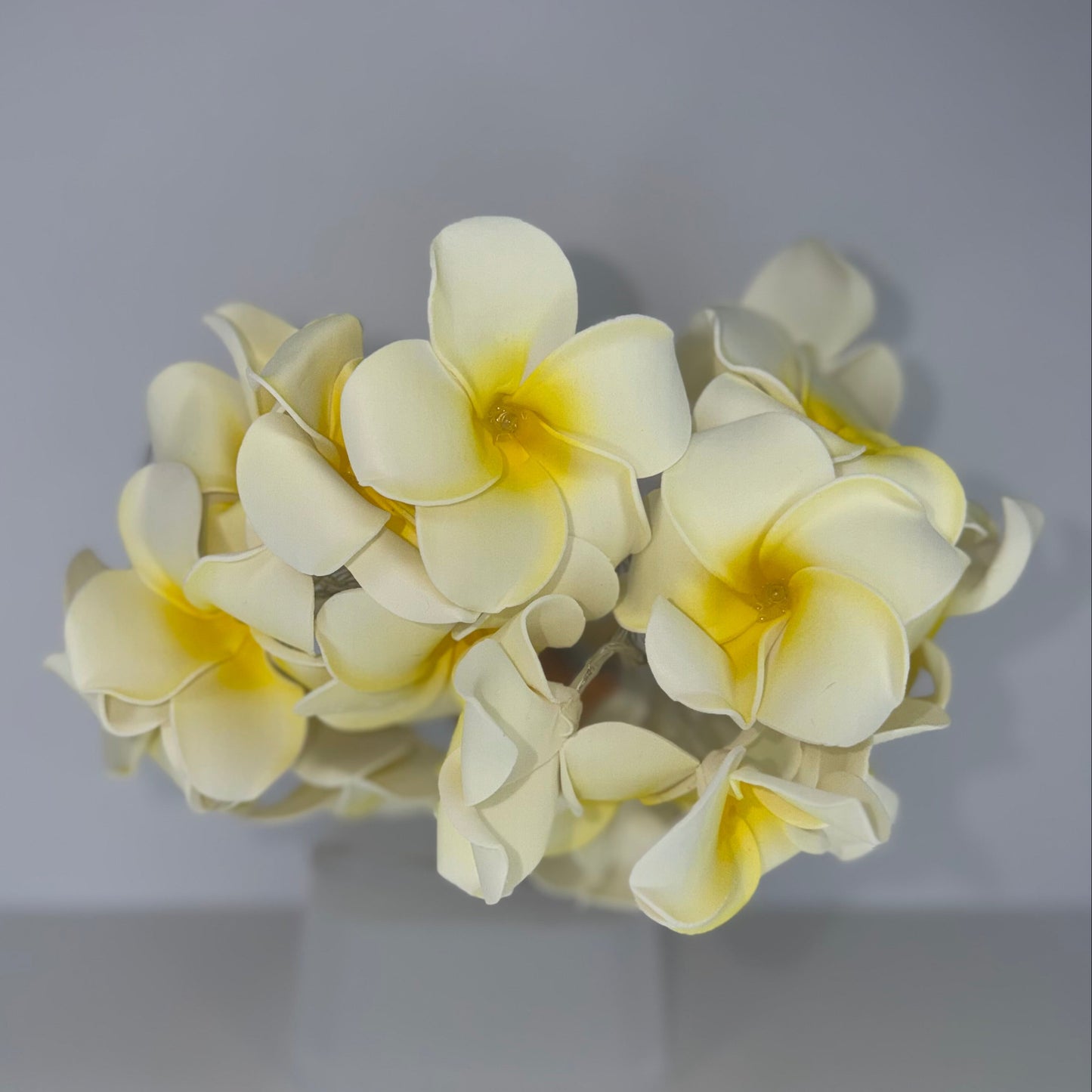 A cluster of artificial plumeria flowers in pale cream with bright yellow centers, displayed on a clear stand against a neutral gray background.