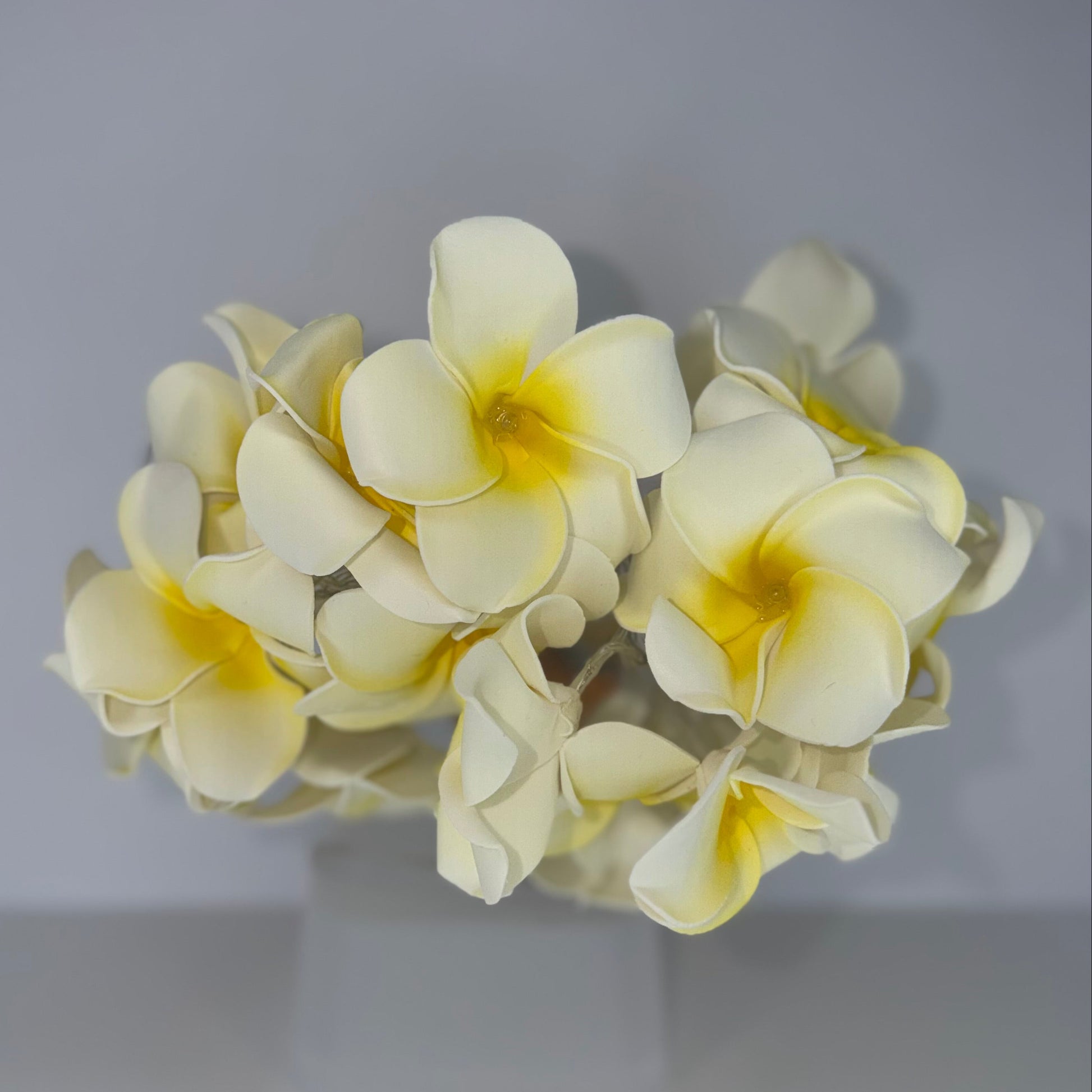 A cluster of artificial plumeria flowers in pale cream with bright yellow centers, displayed on a clear stand against a neutral gray background.