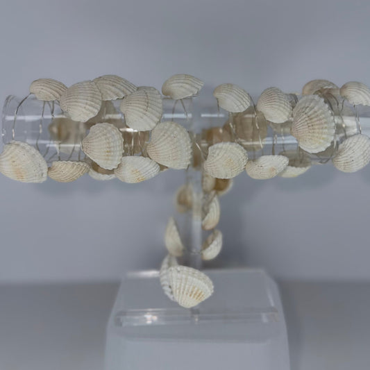 A decorative T-shaped wall light fixture made from numerous small white and beige seashells attached to clear acrylic tubes, mounted on a translucent white plastic base against a plain light gray background.