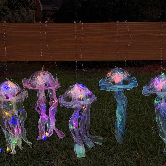 Five translucent jellyfish-shaped hanging ornaments with internal LED fairy lights in pink, blue, purple, green, and yellow, displayed outdoors on grass in front of a wooden fence at night.