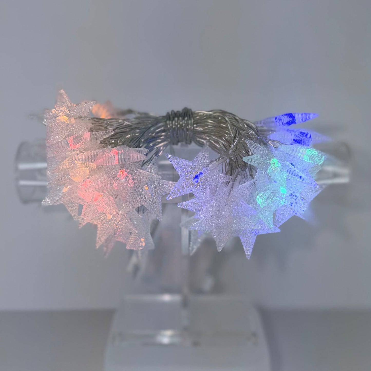 Star-shaped string lights glowing in multiple colors, including pink, blue, and white, displayed on a clear acrylic stand against a neutral gray background.
