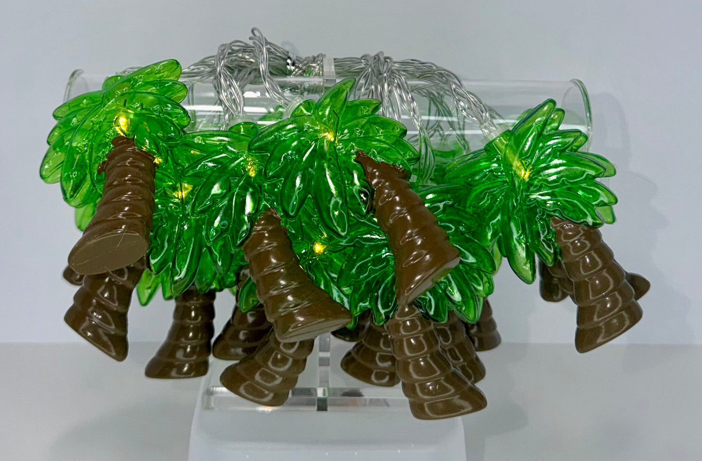 A decorative display stand holding a clear cylindrical tube filled with green translucent palm-leaf shapes, brown boot-like shapes, and a coiled silver wire with small glowing lights, set against a plain light background.