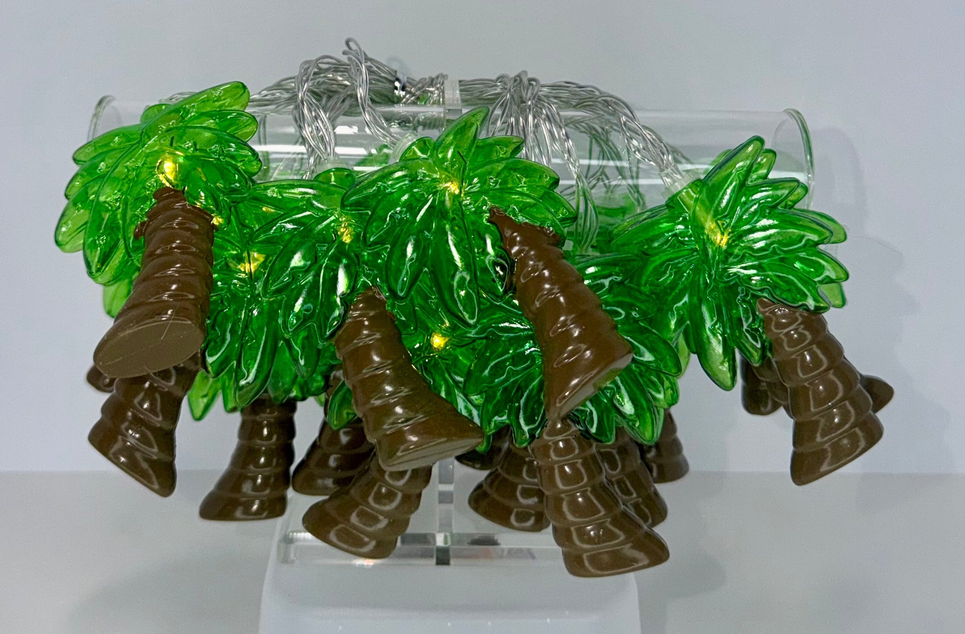 A decorative display stand holding a clear cylindrical tube filled with green translucent palm-leaf shapes, brown boot-like shapes, and a coiled silver wire with small glowing lights, set against a plain light background.