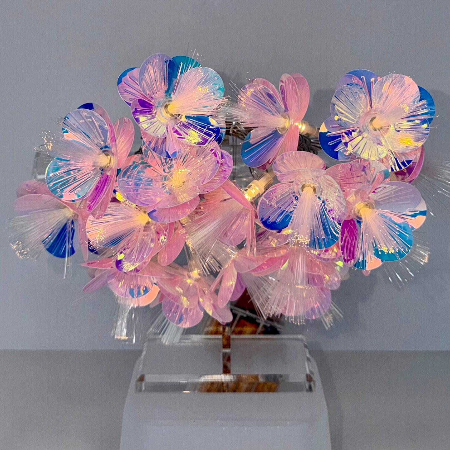 A decorative floral arrangement made from translucent iridescent plastic petals on a clear acrylic stand, photographed against a plain light gray wall and white surface.