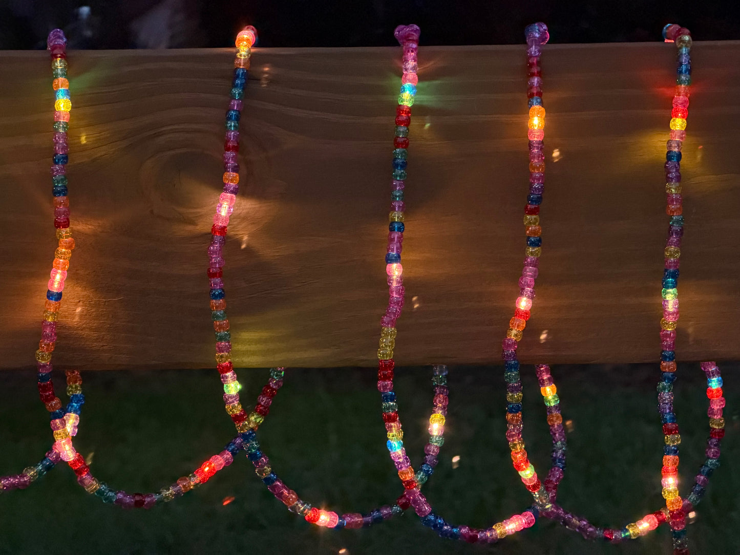 Multicolor LED beaded string lights draped over a wooden fence rail at night, casting warm and cool colored glows onto the wood and grass below.