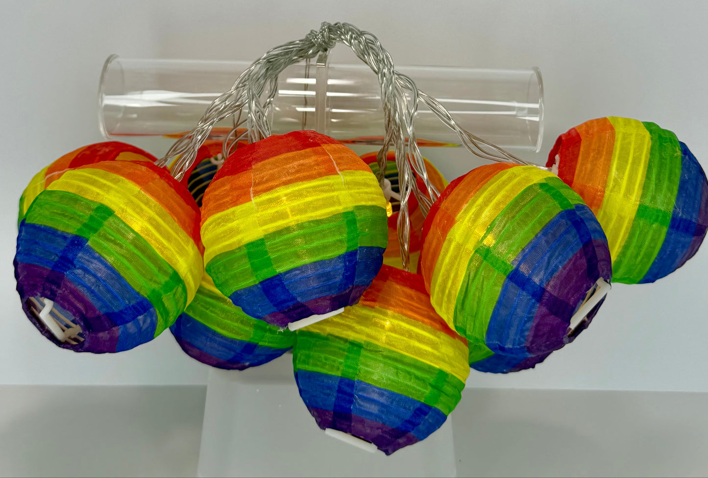 A set of small rainbow paper lanterns with a silver string, displayed on a clear cylindrical stand against a light gray background.