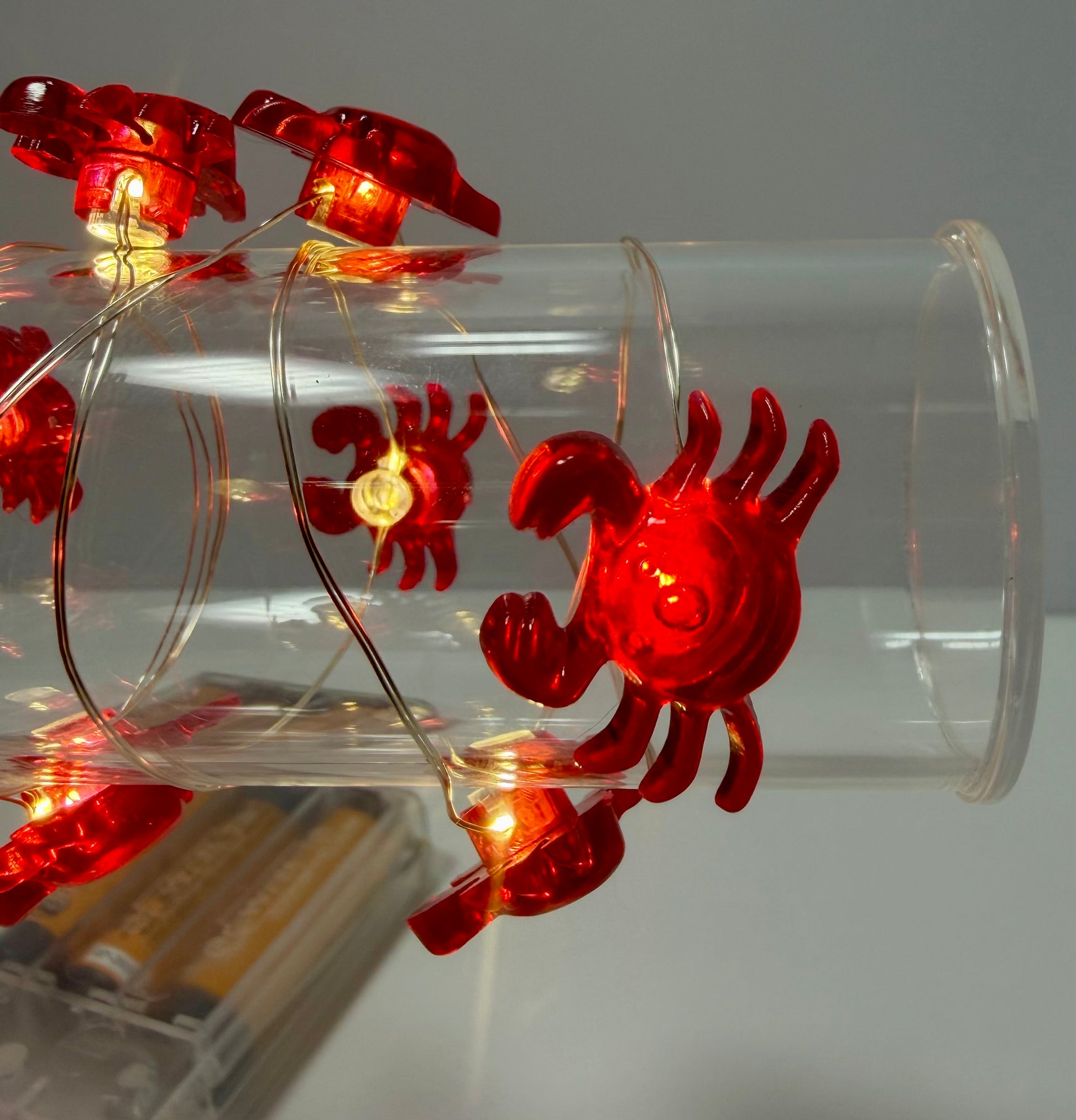 Red crab-shaped string lights glowing with warm white LEDs, draped over a clear plastic tube. The lights are shown close-up against a neutral gray background, with a battery pack partially visible in the lower-left corner.