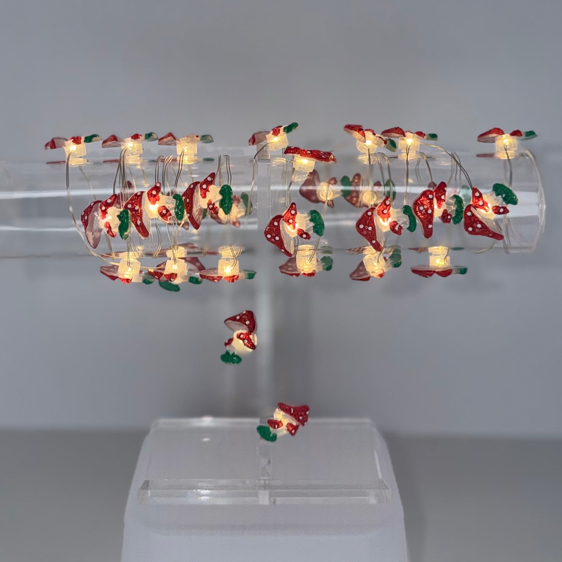 A clear acrylic holiday light display stand holding multiple small LED Christmas lights with warm white bulbs and red, green, and gold ornaments, mounted on a white plastic base against a plain gray studio background.