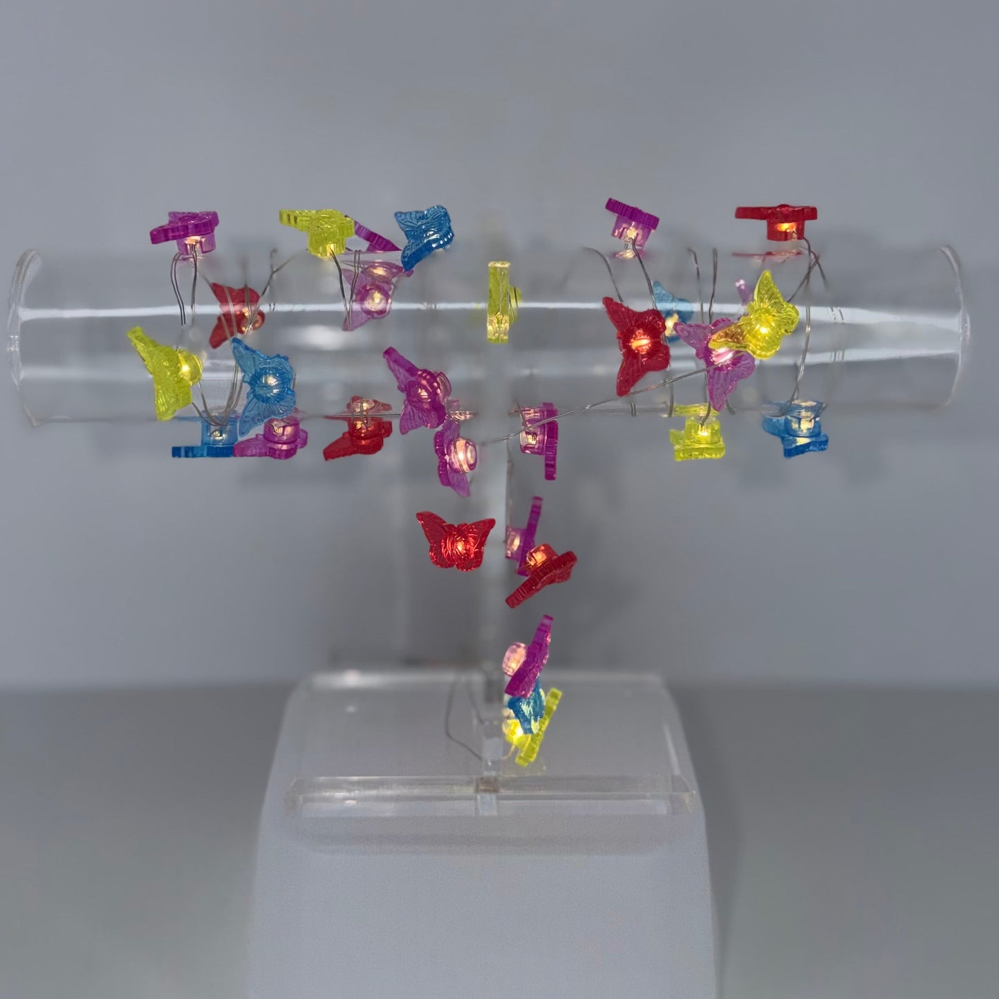 A clear acrylic display stand holding multiple small colorful butterfly-shaped clips or pins in red, yellow, blue, and purple, set against a plain light gray background.
