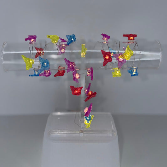 A clear acrylic display stand holding multiple small colorful butterfly-shaped clips or pins in red, yellow, blue, and purple, set against a plain light gray background.