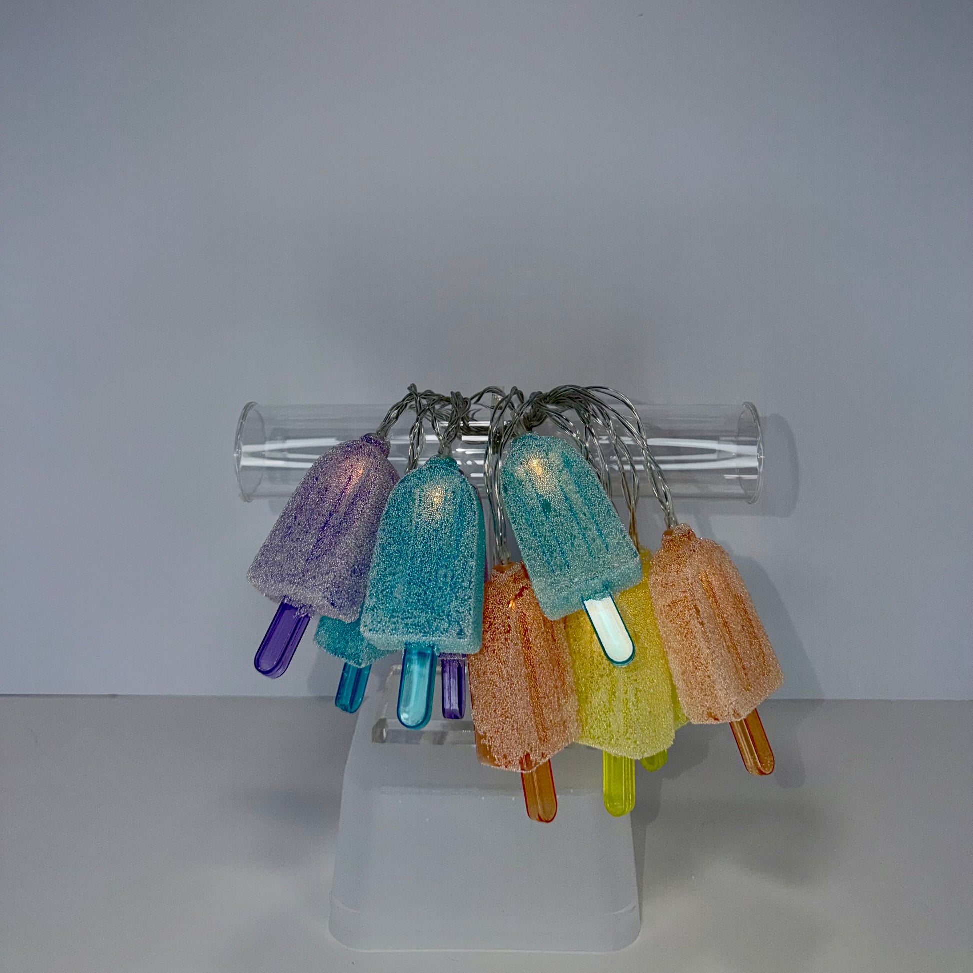 A set of colorful, textured popsicle-shaped string lights hanging from a clear cylindrical rod on a translucent acrylic stand against a plain light gray background.