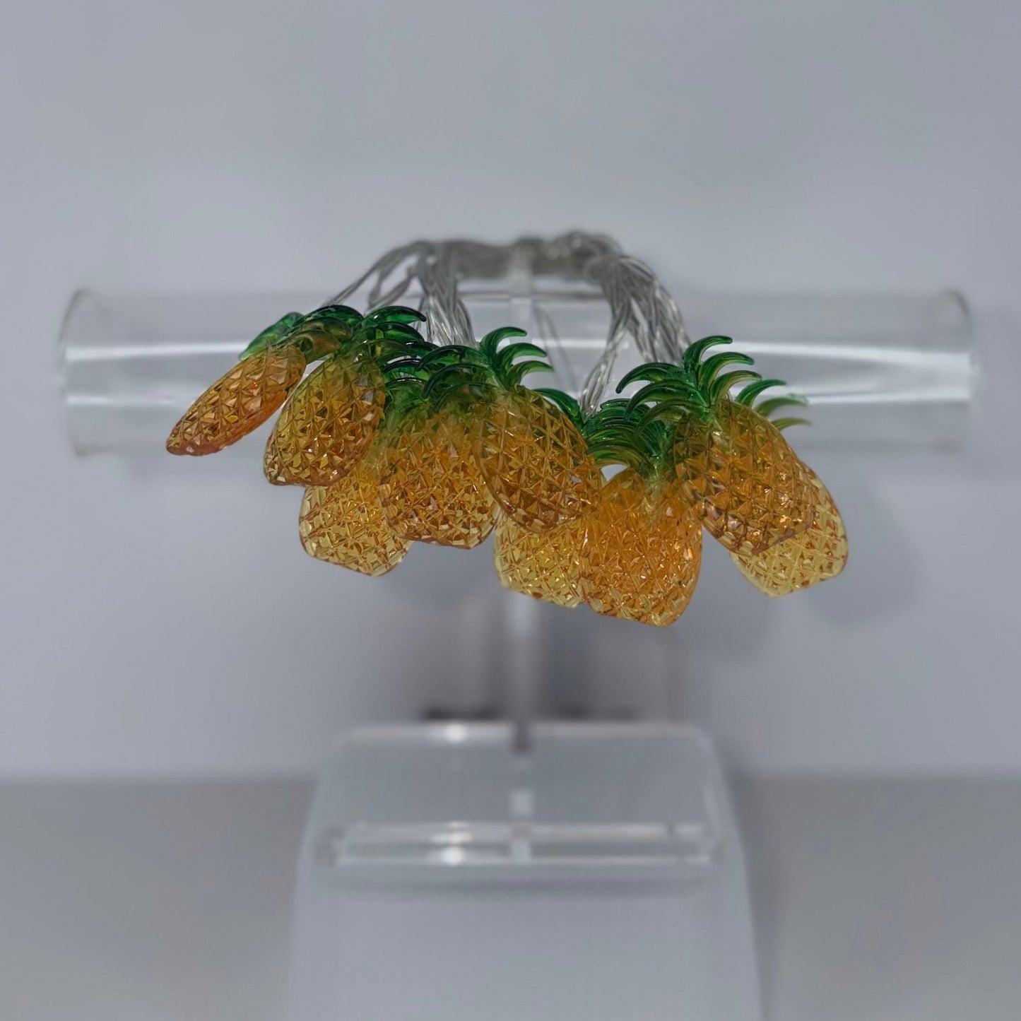 A pair of silver hoop earrings decorated with three yellow textured pineapple charms and green leafy tops, displayed on a clear acrylic stand against a neutral gray background.