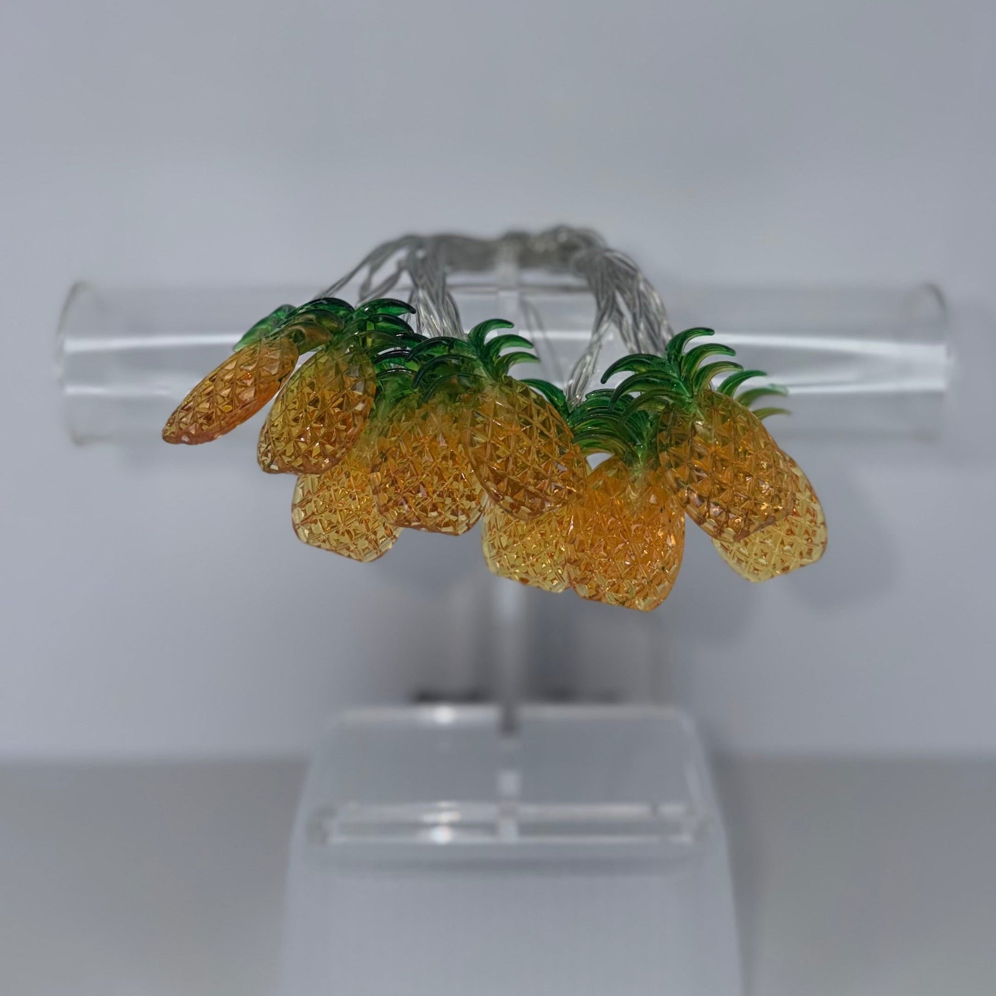 A pair of silver hoop earrings decorated with three yellow textured pineapple charms and green leafy tops, displayed on a clear acrylic stand against a neutral gray background.