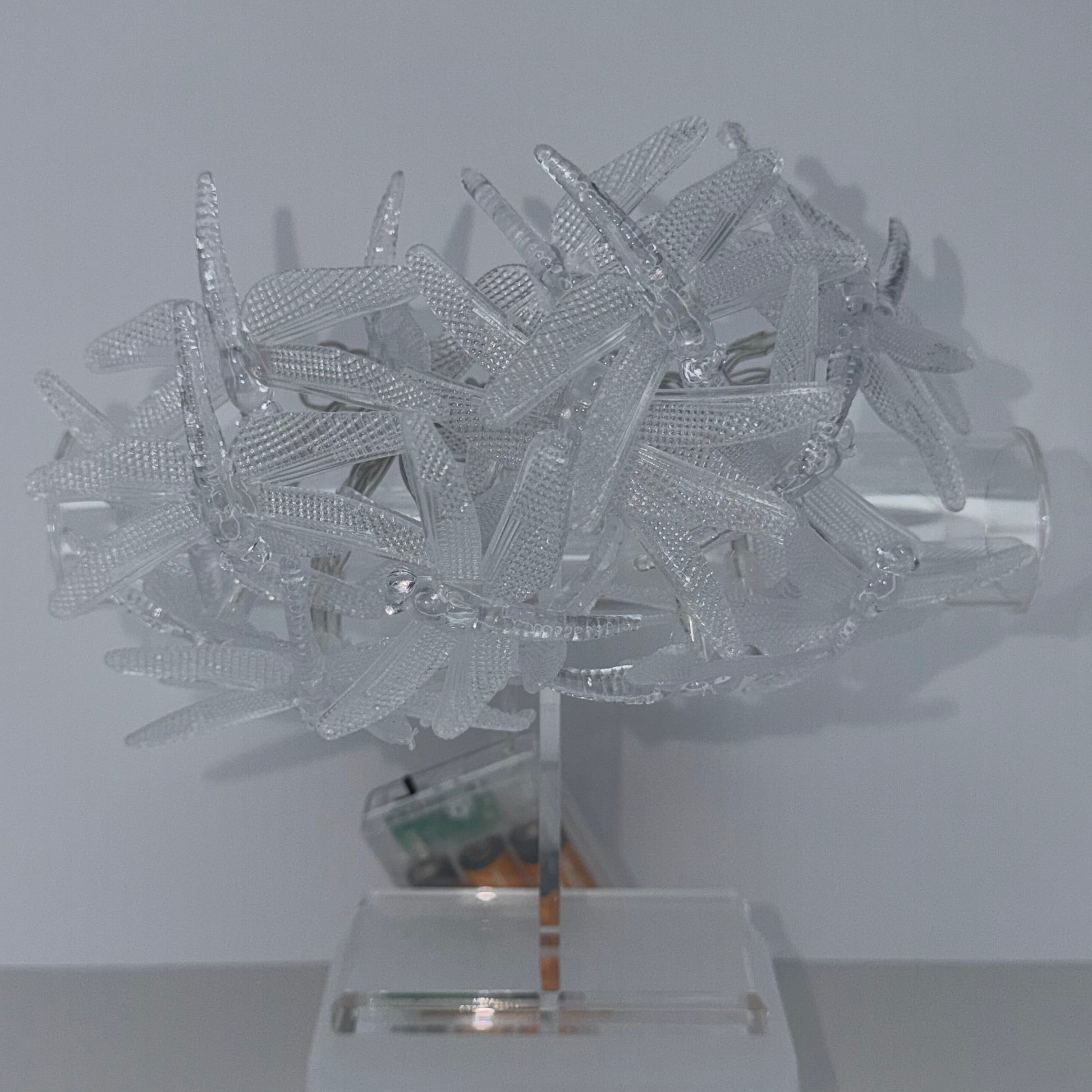 A modern wall-mounted light fixture with a cluster of clear ribbed plastic or acrylic strips forming the shade, attached to a white square wall plate on a plain light gray wall.