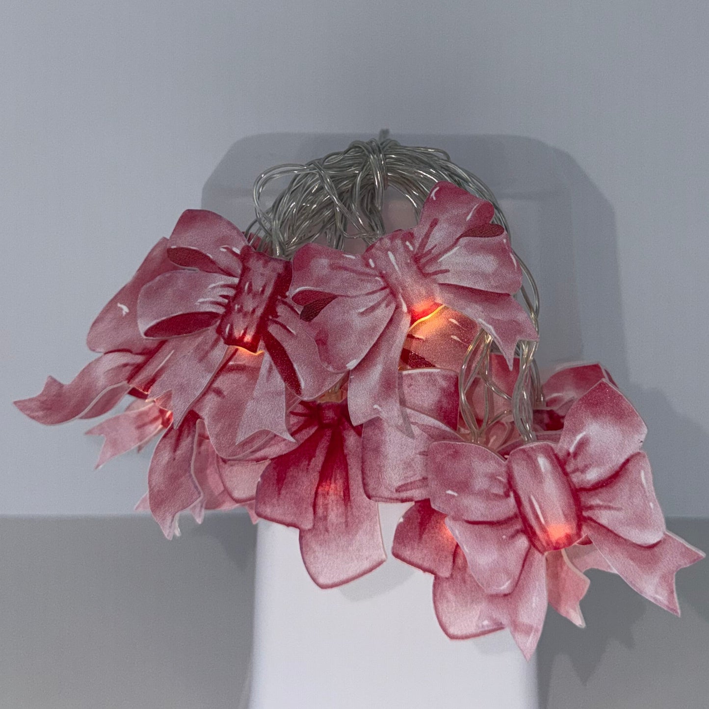 A pink silk flower arrangement with a warm orange glow from hidden fairy lights, displayed on a white rectangular block against a minimalist gray background.