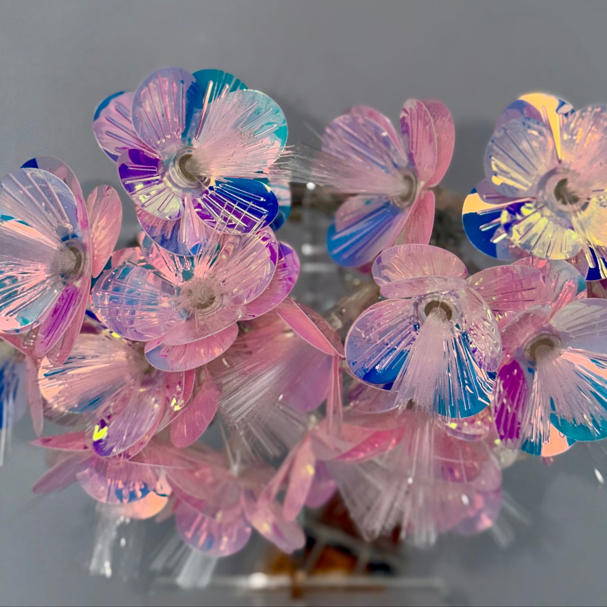 A cluster of iridescent plastic flower-shaped ornaments or charms in pastel pink, blue, purple, and yellow tones, photographed against a plain gray background.