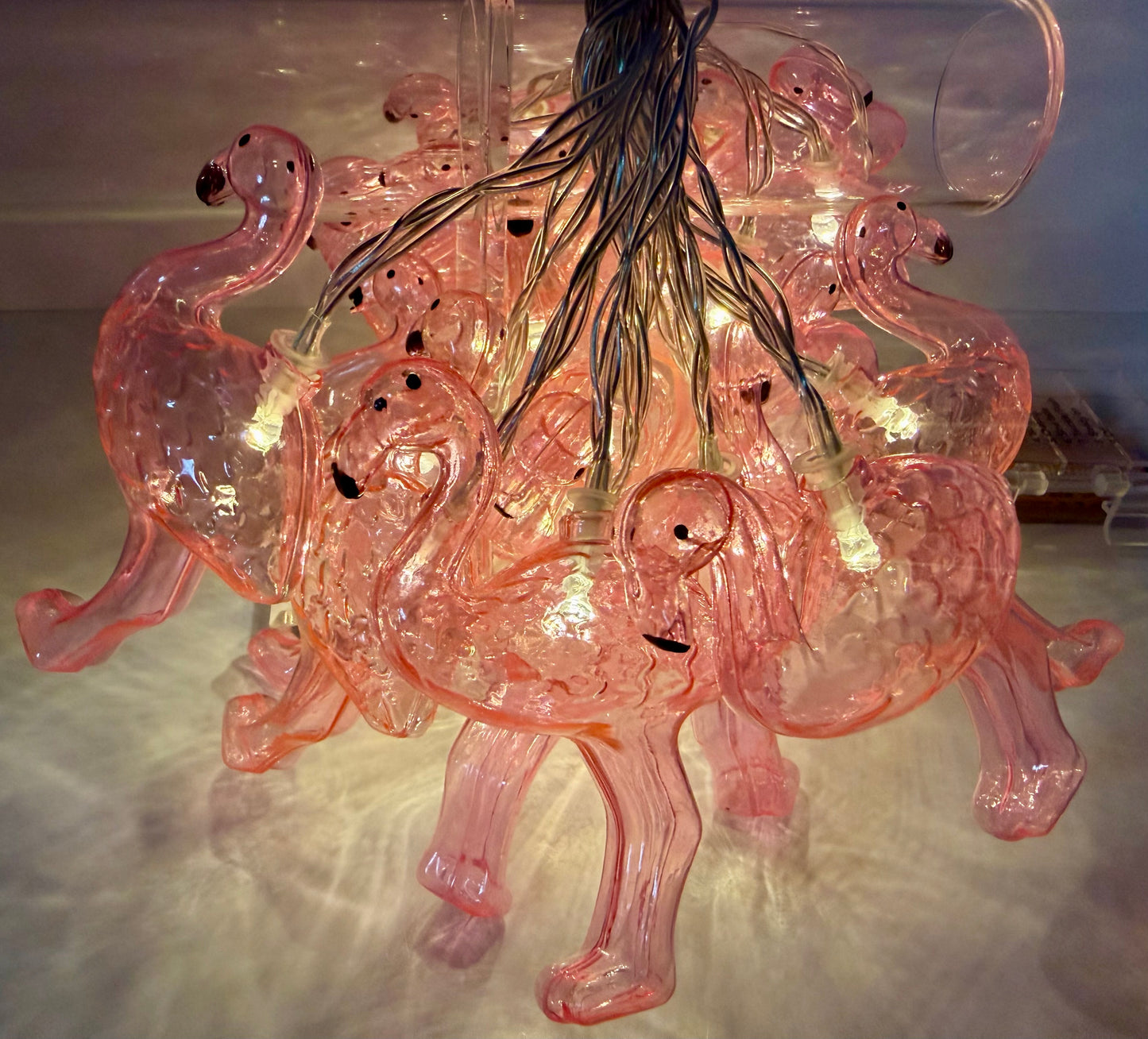 A pink glass chandelier featuring multiple flamingo-shaped bulbs with warm white string lights, photographed against a neutral indoor background.
