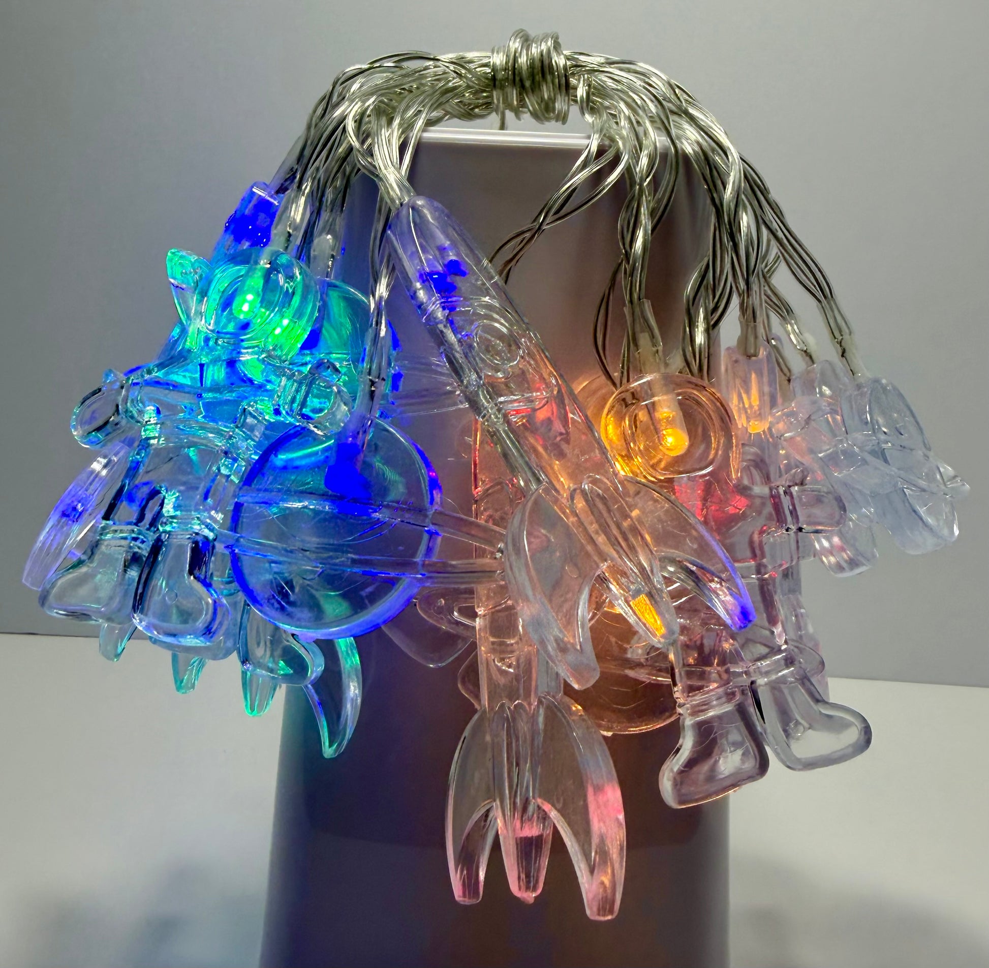 Multicolor LED fairy string lights with clear plastic clip-on bulbs and silver wire, shown draped over a dark cylindrical display stand against a neutral gray background.