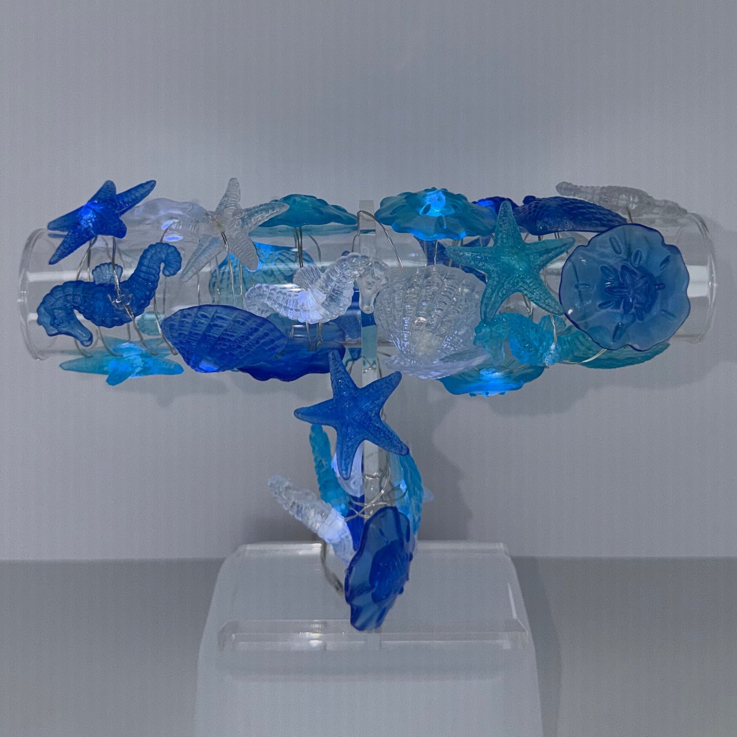 A decorative wall-mounted fixture made from transparent and blue glass-like elements, shaped like a stylized figure with starfish details, attached to a white wall on the right against a gray corner background.
