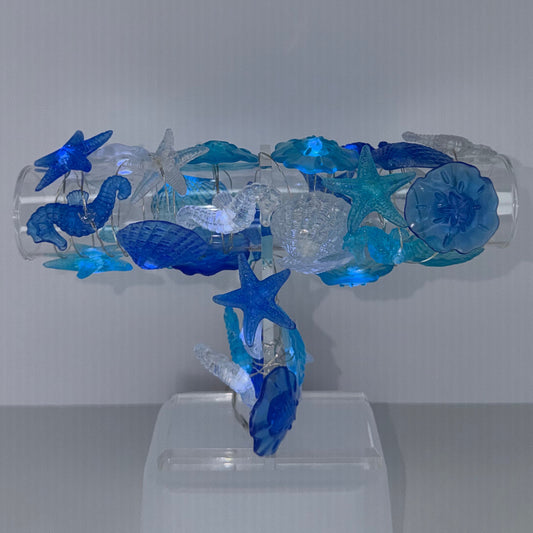A decorative wall-mounted fixture made from transparent and blue glass-like elements, shaped like a stylized figure with starfish details, attached to a white wall on the right against a gray corner background.