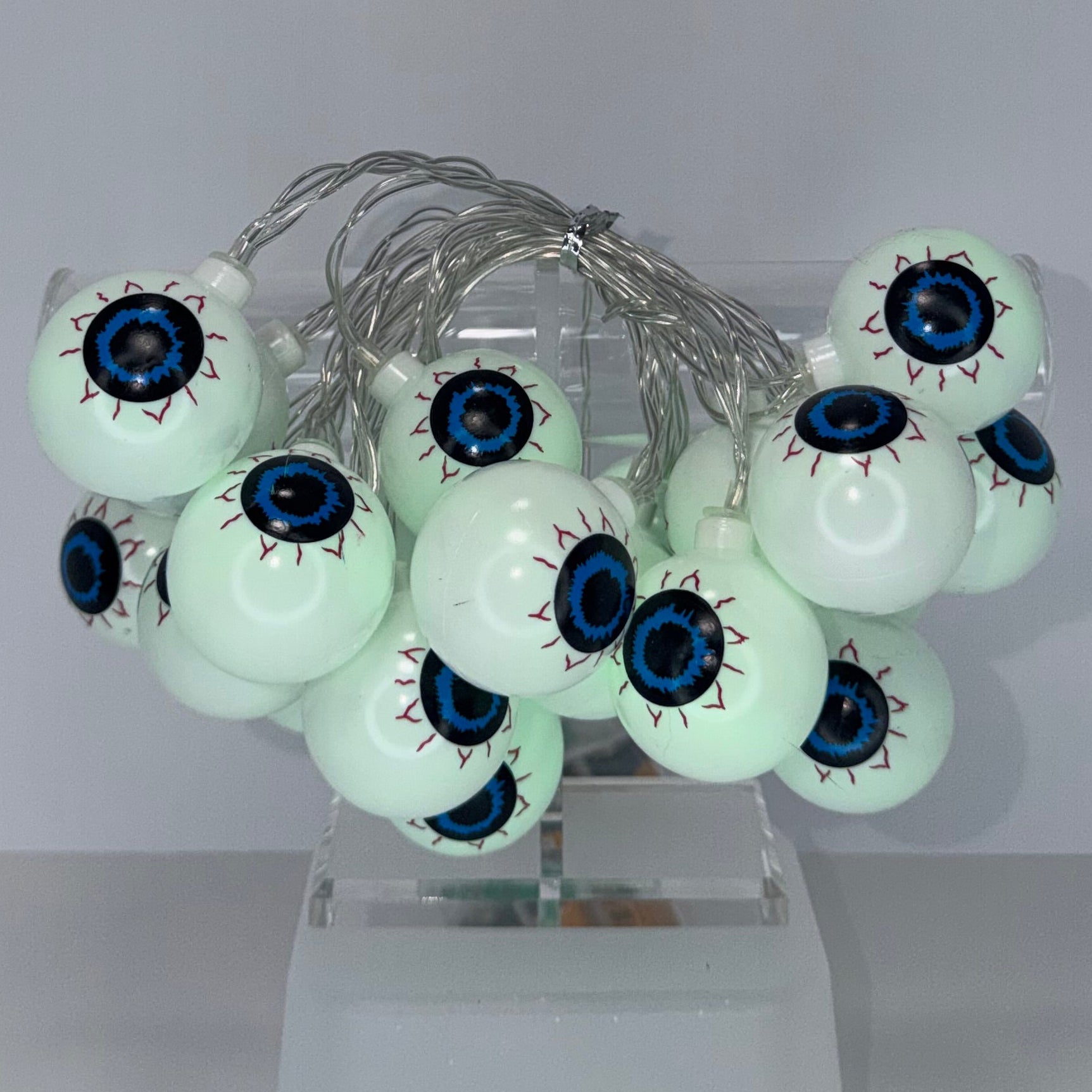 Glowing white eyeball string lights with realistic blue and black irises, wrapped in silver wire and shown plugged into a white wall outlet against a plain white wall.