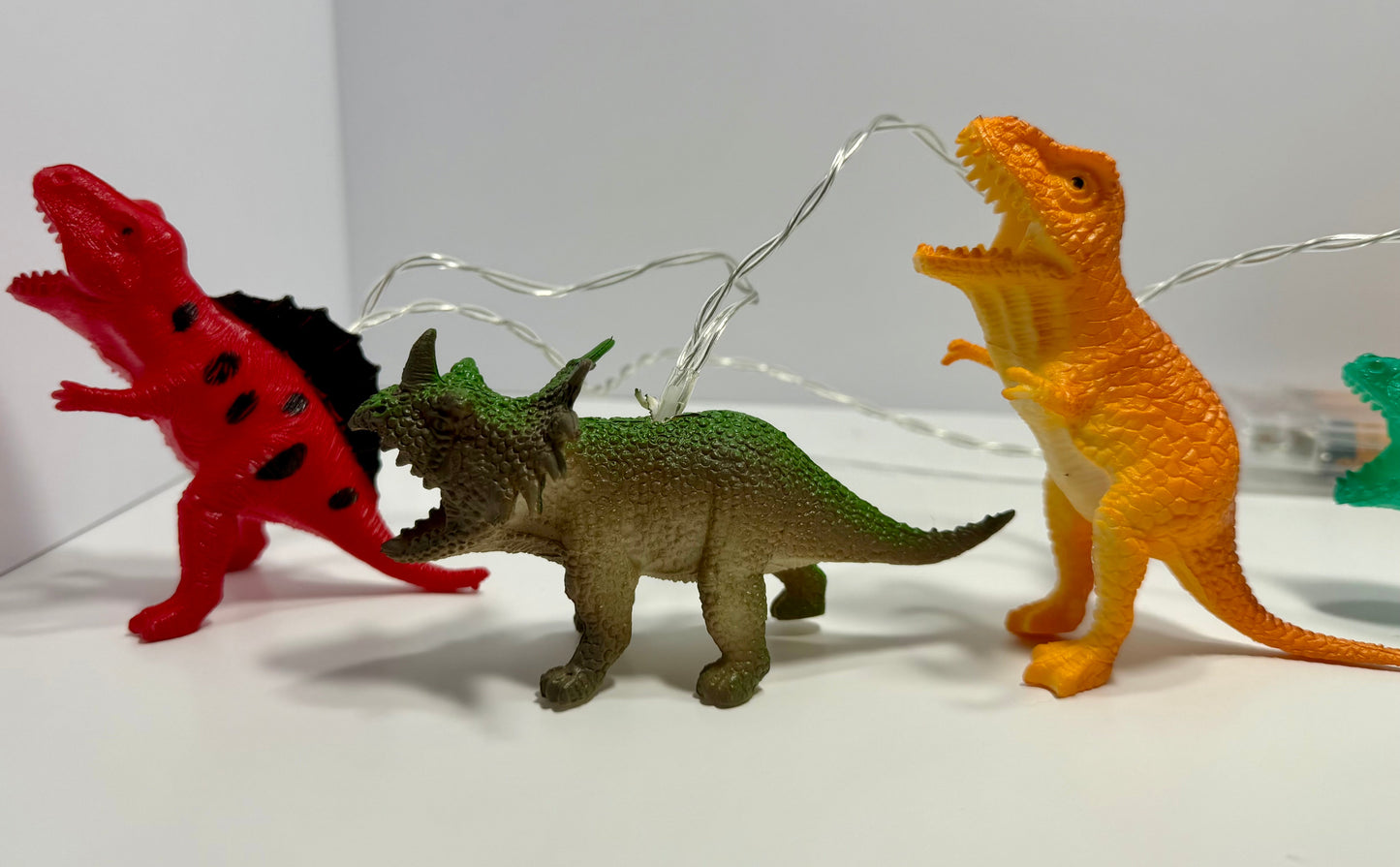 Three colorful dinosaur toy figurines arranged on a white surface against a white background, with thin string lights draped behind them. The left dinosaur is red with black spots and a black back, the center dinosaur is green and brown, and the right dinosaur is orange-yellow.