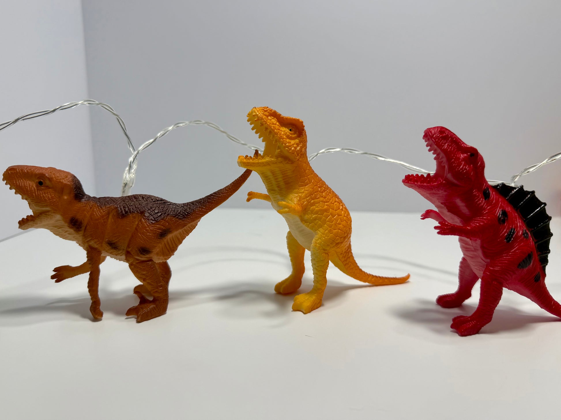 Three small dinosaur toy figurines in brown, yellow-orange, and red, arranged in a row on a white surface with thin silver string lights behind them against a plain light gray background.