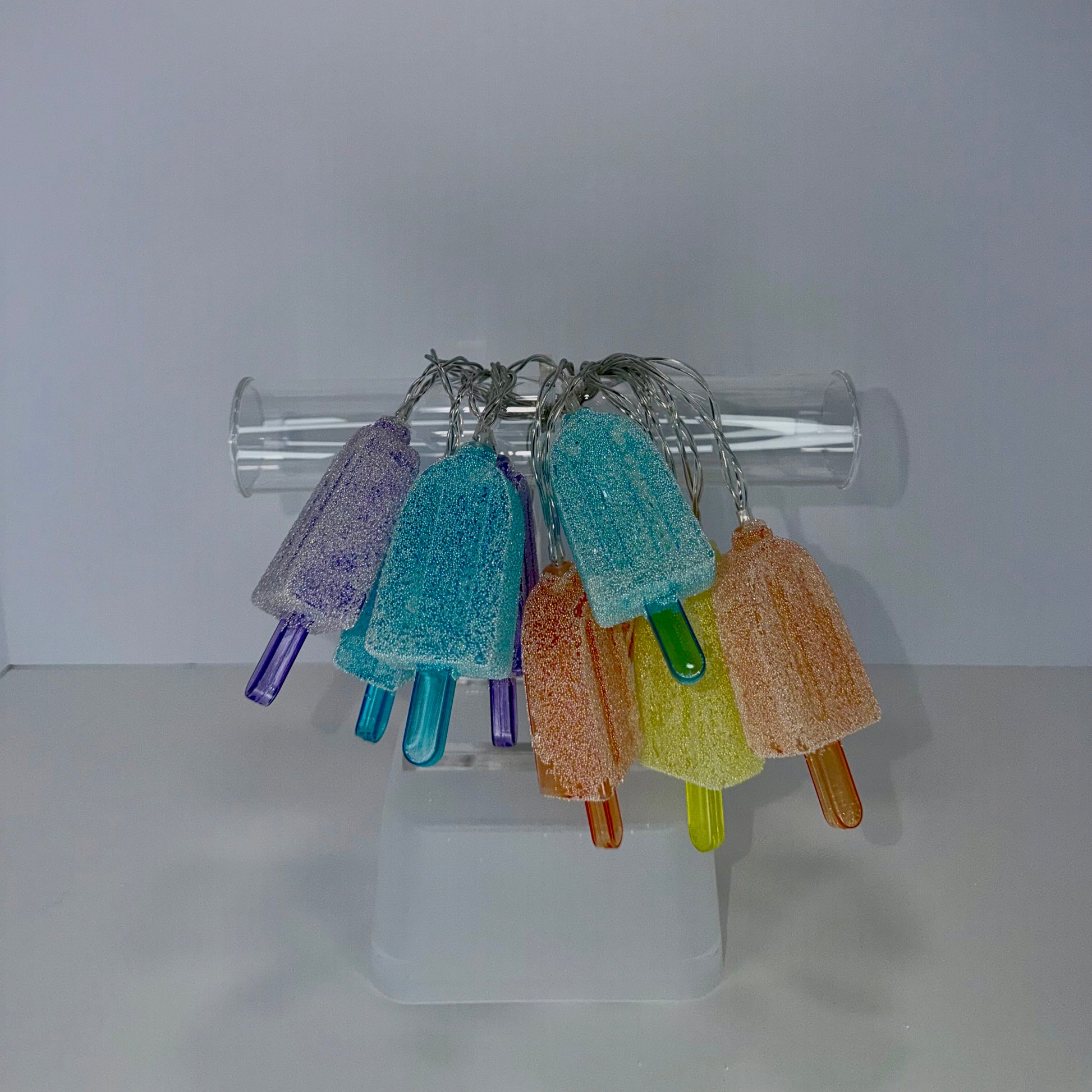 A set of colorful, sugar-coated popsicle-shaped keychains hanging from a clear plastic tube stand on a white surface against a light gray background.