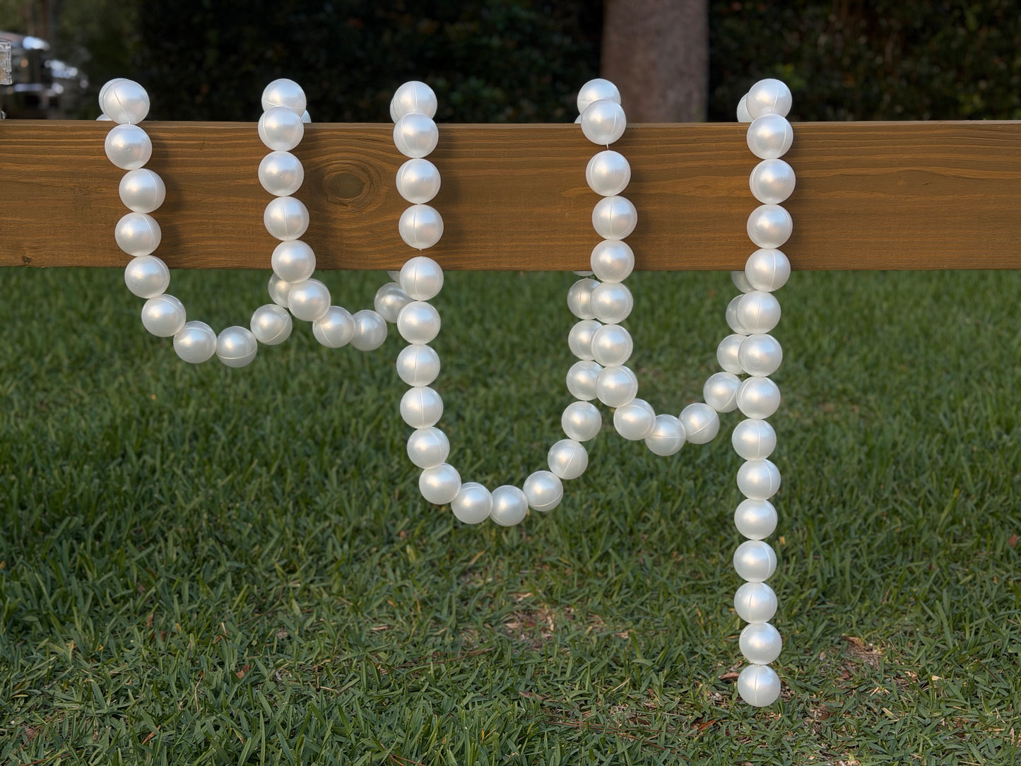 White faux-pearl letter garlands spelling 'W' and 'M' draped over a wooden fence on a green grass lawn.