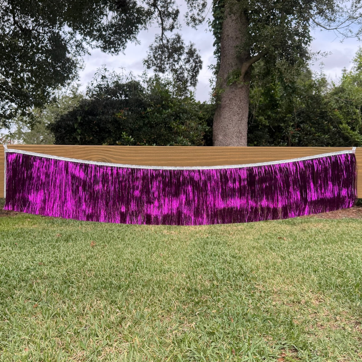 A bright purple metallic tinsel fringe banner draped across an outdoor grassy lawn with trees in the background.
