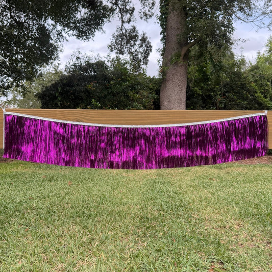 A bright purple metallic tinsel fringe banner draped across an outdoor grassy lawn with trees in the background.