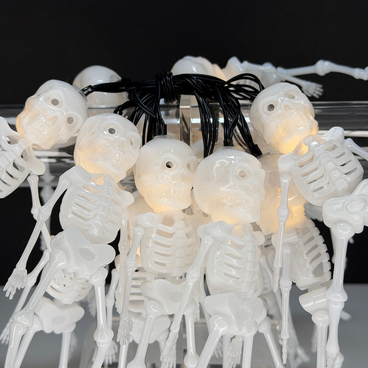 A string of white plastic skeleton bone lights with glowing warm-white LED bulbs inside each skull, connected by a black cord, shown against a dark background.