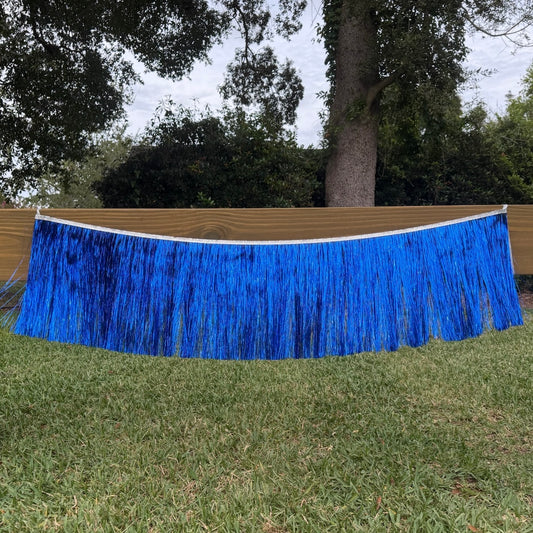 A bright blue tinsel fringe banner hanging outdoors on a wooden fence above a green lawn, with trees in the background.