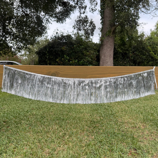 A silver metallic fringe banner or backdrop hanging outdoors over a wooden surface on a grassy lawn, with trees and bushes in the background.
