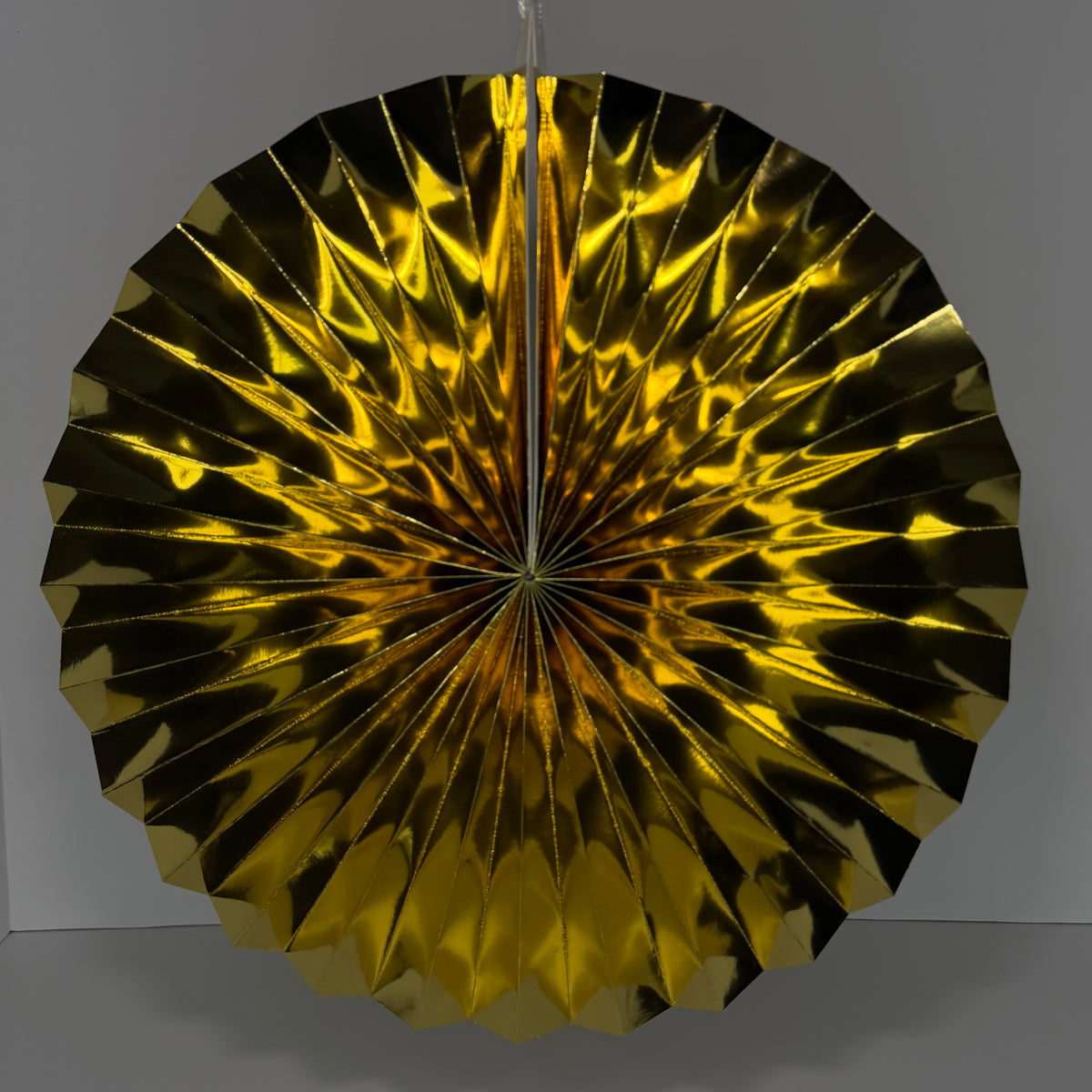 A round, metallic gold paper fan decoration with a pleated, accordion-like design, hanging against a neutral gray background.