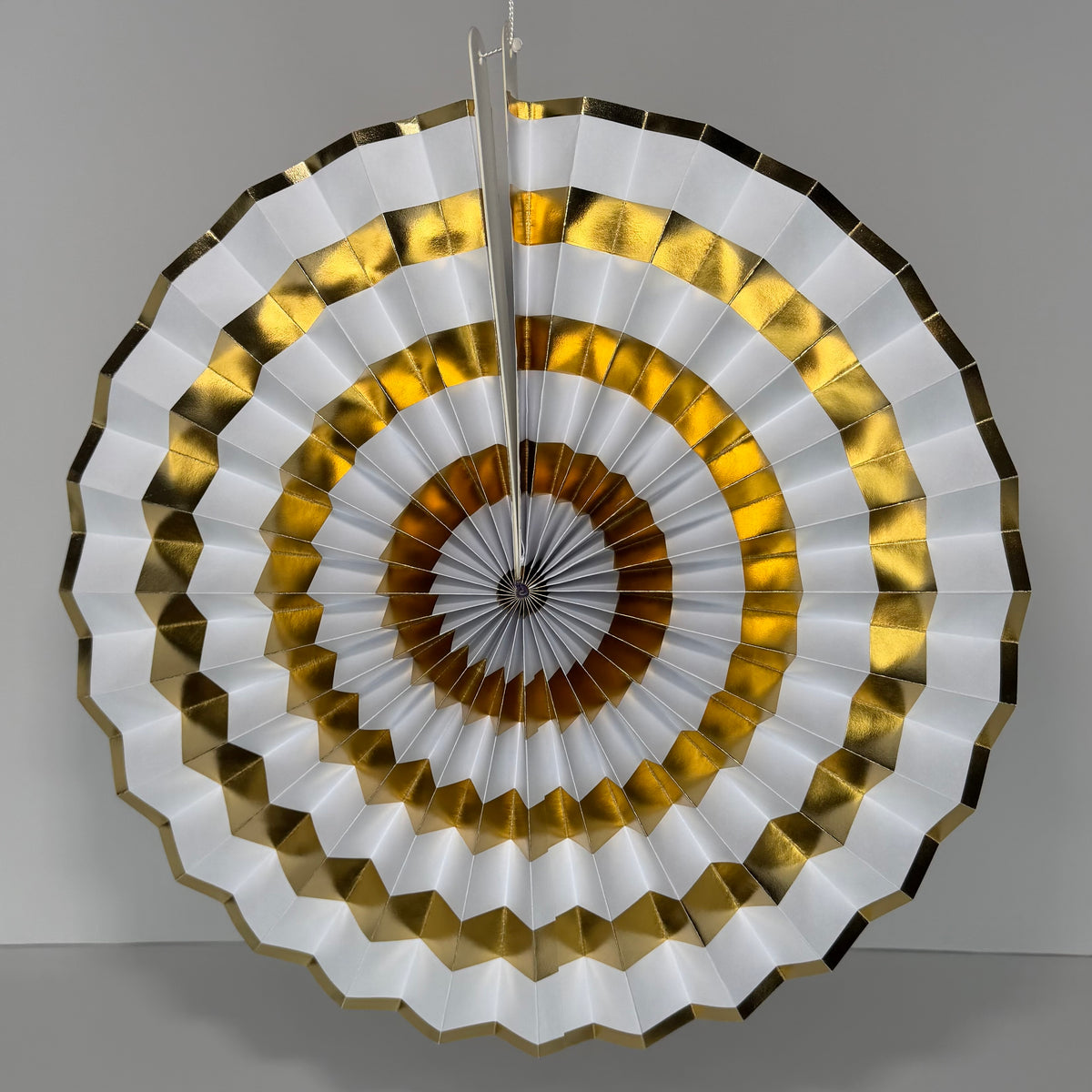 A circular pleated hanging fan or wall decor piece with concentric rings of white, metallic gold, and brown, suspended against a plain light gray wall.