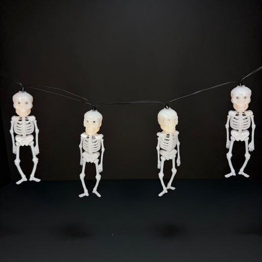 Four hanging plastic skeleton decorations with attached faces, suspended on a black string against a dark background.