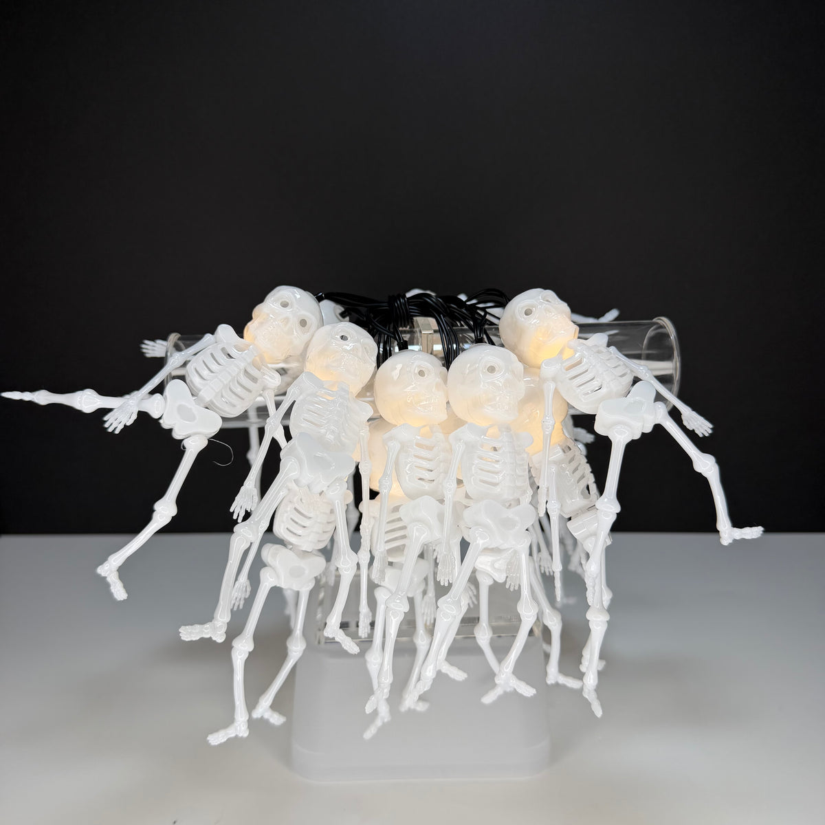 A cluster of small white plastic skeleton figures posed together on a clear cylindrical stand, with warm white string lights glowing inside their heads. The background is a dark black-to-gray gradient with a light gray surface below.