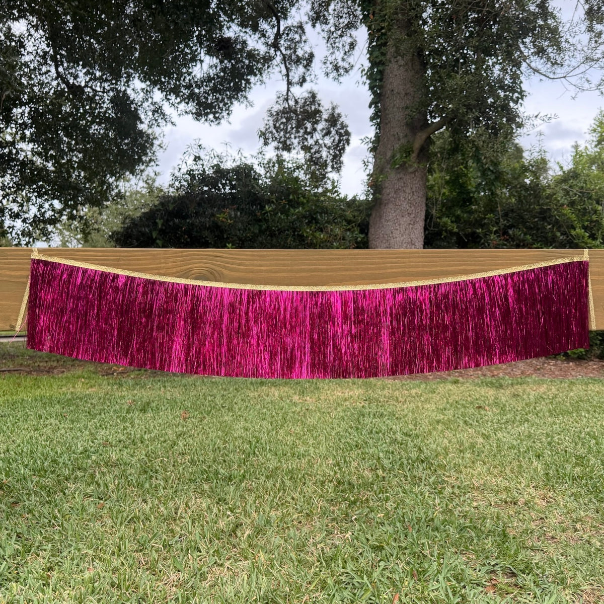 A bright magenta metallic fringe banner draped across a wooden fence in an outdoor backyard setting with green grass and trees in the background.