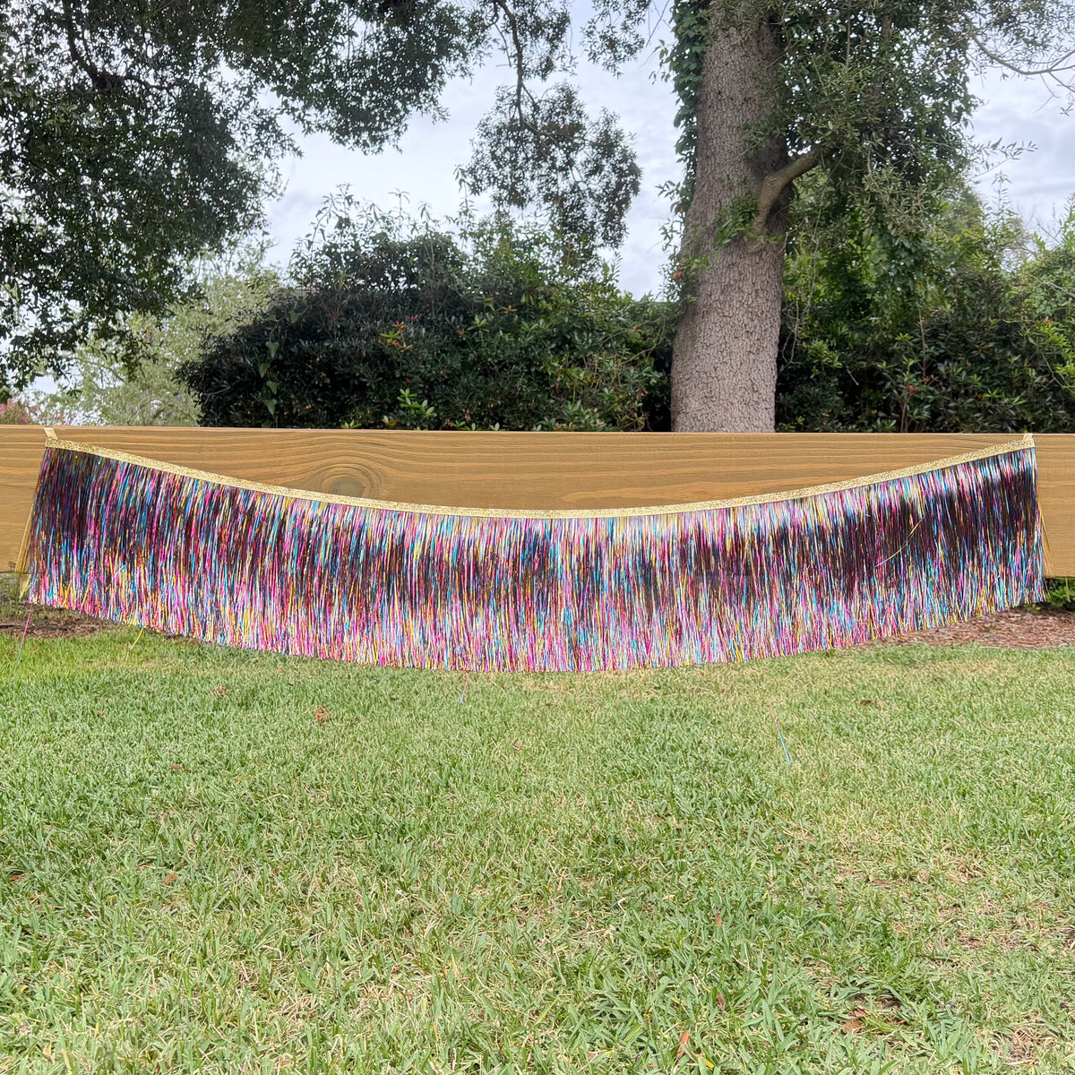 A long, wide fringe banner with multicolored strands hanging horizontally, displayed outdoors on a wooden fence above a green lawn with trees in the background.