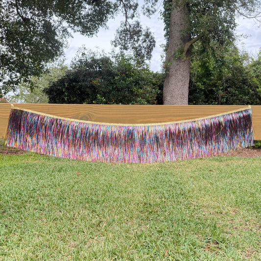 A long, wide fringe banner with multicolored strands hanging horizontally, displayed outdoors on a wooden fence above a green lawn with trees in the background.