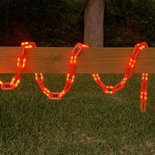 Red LED chain link string lights draped over a wooden fence rail at night, with a grassy yard and tree trunk in the background.