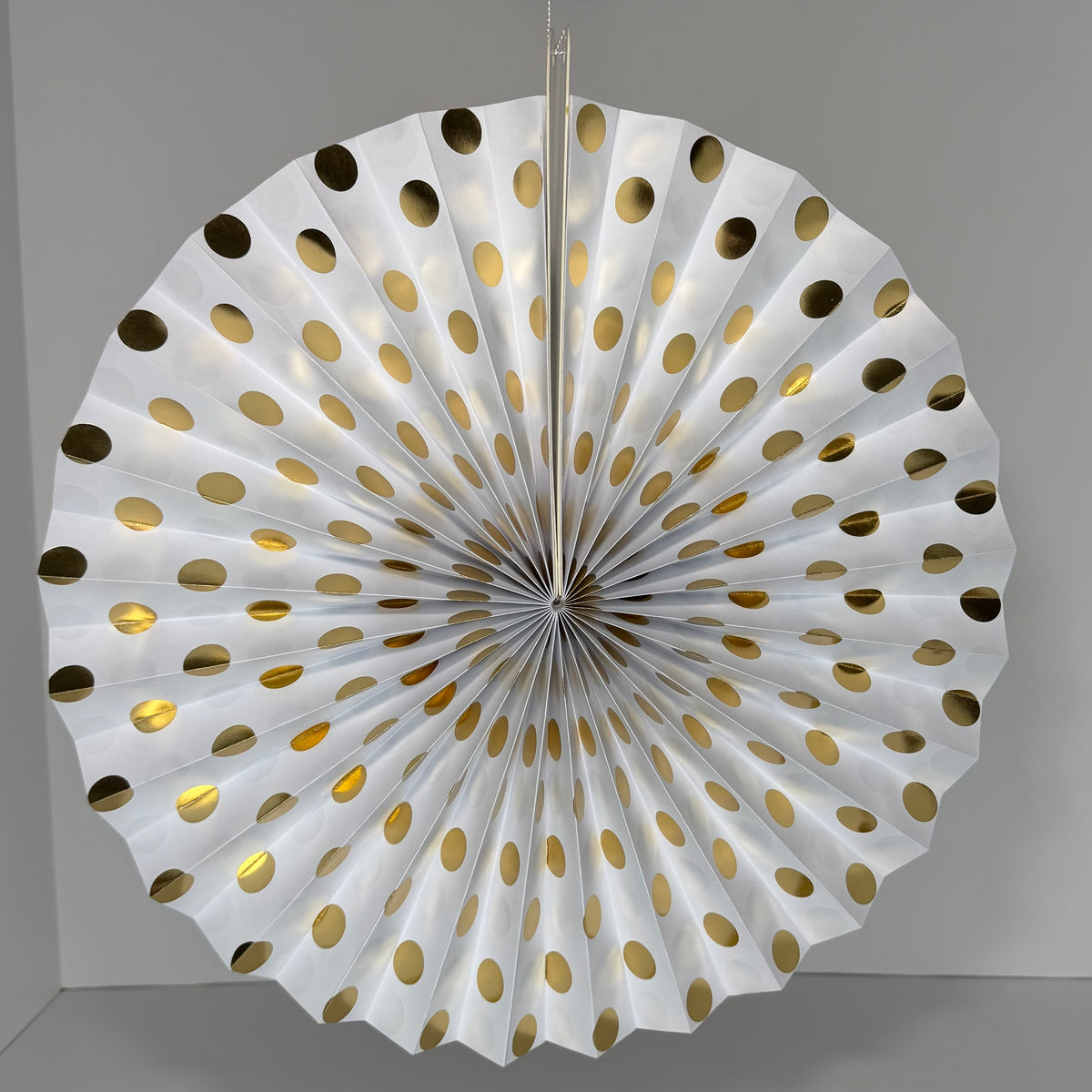 A white paper fan decoration with metallic gold polka dots, hanging against a neutral gray background.