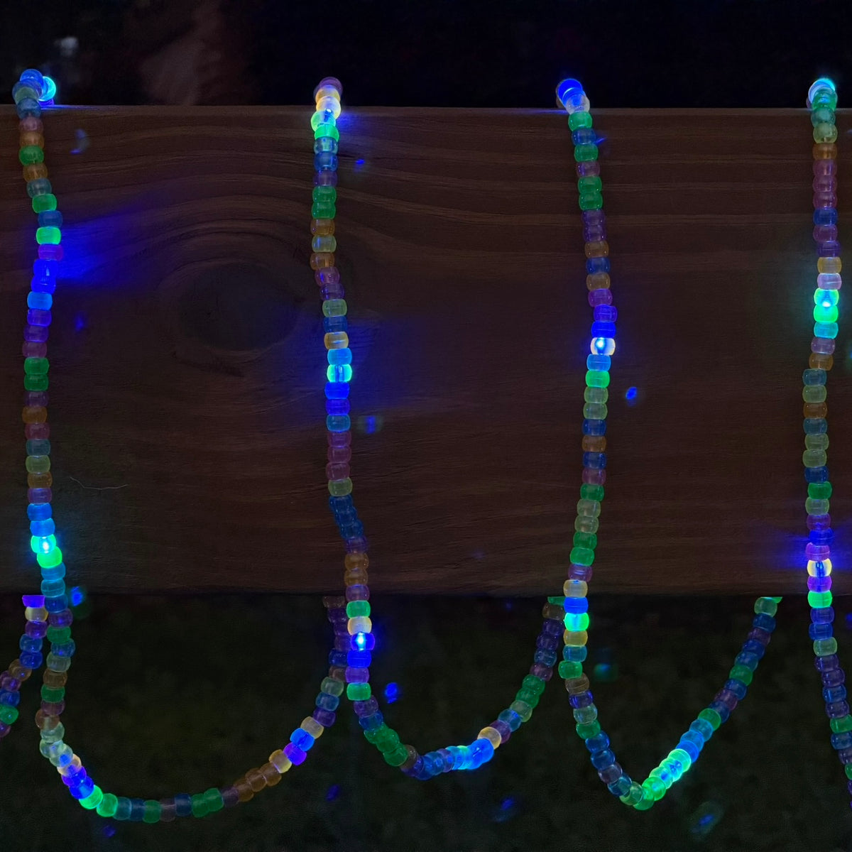 Multicolor LED beaded string lights draped over a wooden fence in a dark outdoor setting, glowing with blue, green, orange, and purple lights.