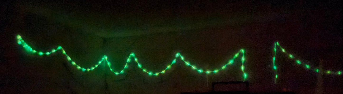 Green string lights draped across a dark room corner, with a small illuminated box or cabinet below on a very dark background.