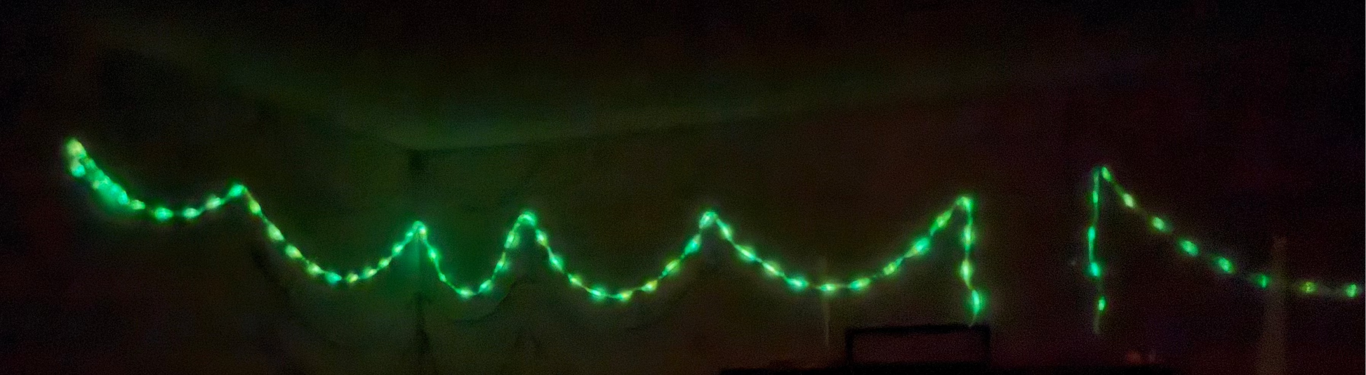 Green string lights draped across a dark room corner, with a small illuminated box or cabinet below on a very dark background.