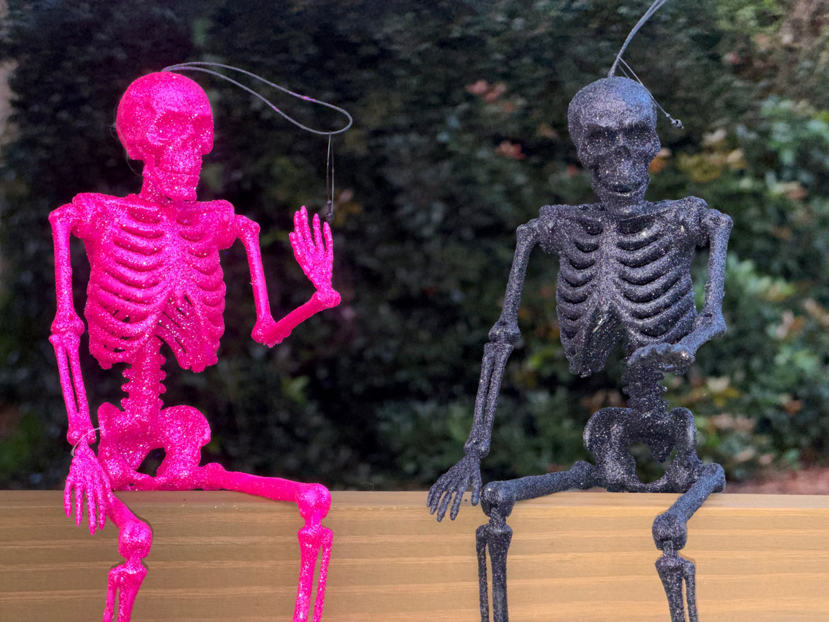 Hot pink and black glitter skeletons posed sitting on a center rail waving in the daytime