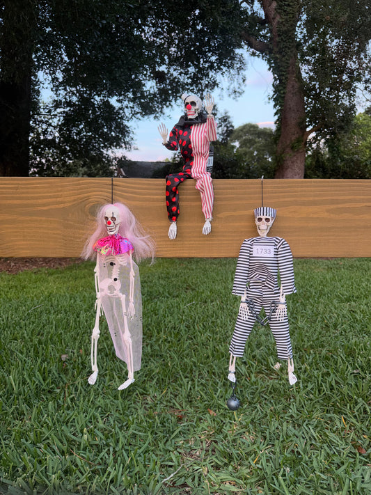 Three Halloween yard decorations hanging on a wooden fence in a backyard setting. The set includes a hanging skeleton clown with pink hair and a pink collar, a hanging skeleton prisoner in a black and white striped jumpsuit, and a hanging skeleton clown in a red and white striped outfit sitting on the fence.