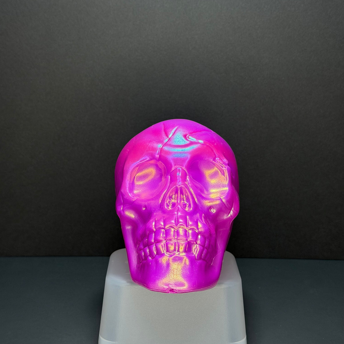 A glossy, iridescent skull-shaped object with a vibrant pink and purple holographic finish, displayed on a white pedestal against a dark gray background.